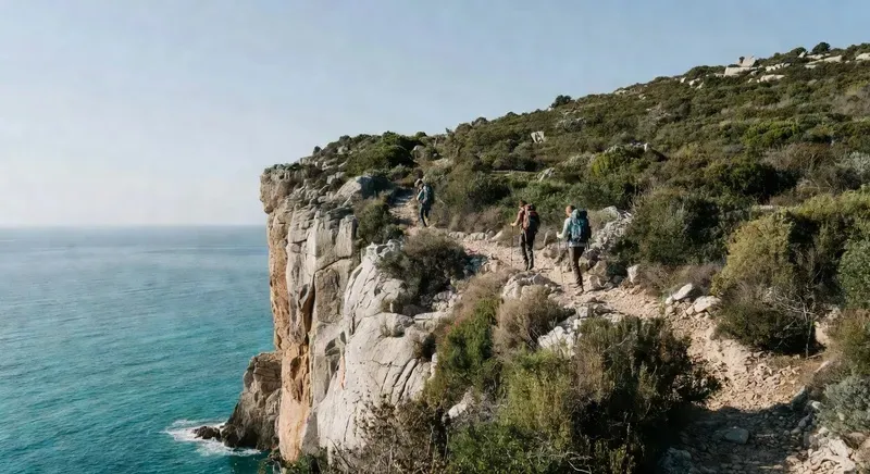Two hikers on a rocky coastal path in Sardinia — narrow trail along limestone cliffs above the turquoise Mediterranean sea