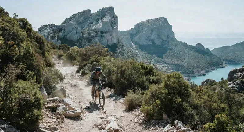 E-bike rider on a mountain trail through the Supramonte with Cala Gonone coastline visible below on a full-day sardinia cycling tour